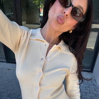 Woman wearing gold hoop earrings, sunglasses, and a cream knit top posing outdoors